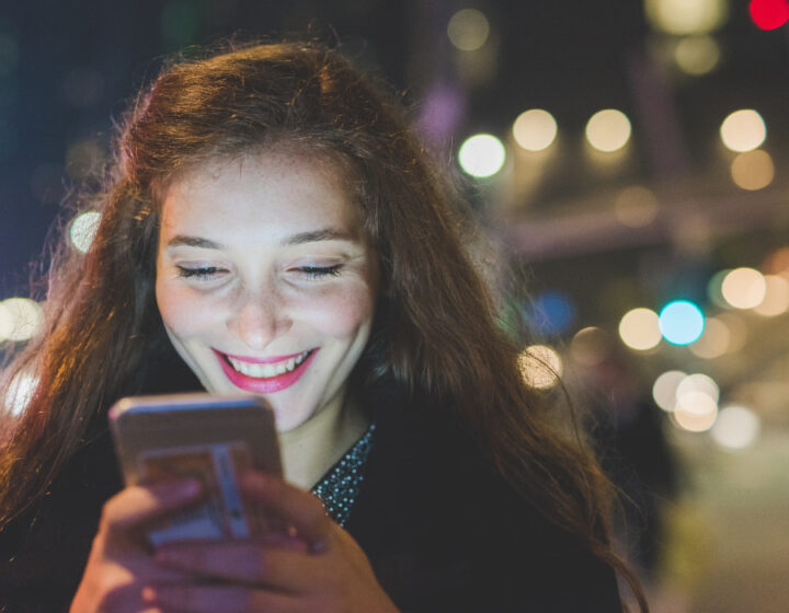 Jonge vrouw kijkt, ’s avonds buiten op straat, lachend naar haar telefoon